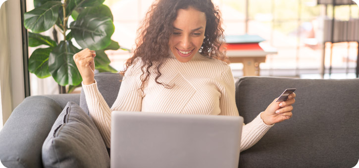 mujer joven feliz con tarjeta de crédito en la mano frente a computadora con buso crema sentada en sillón gris